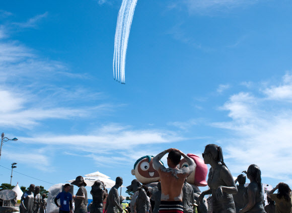‘제16회 보령머드축제’ 개막식이 열린 19일 충남 보령시 신흑동 대천해수욕장 상공에서 공군 특수비행팀 ‘블랙이글스’가 펼치는 에어쇼를  온몸에 머드를 바른 관광객들이 감상하고 있다.  연합뉴스