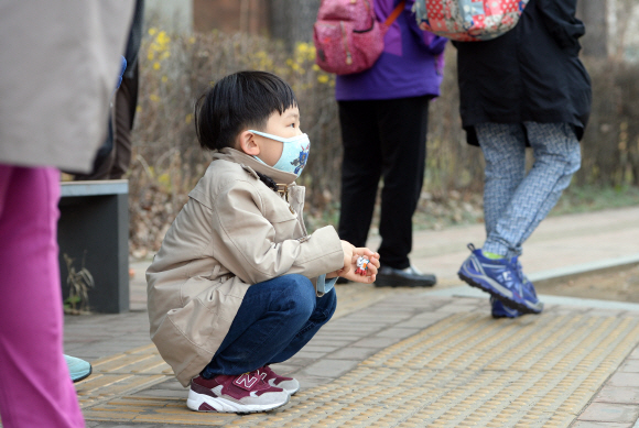 이현이가 집 앞 버스 정류장에서 외할머니와 놀이학교 버스를 기다리며 행인들 사이에 쪼그려 앉아 있다.