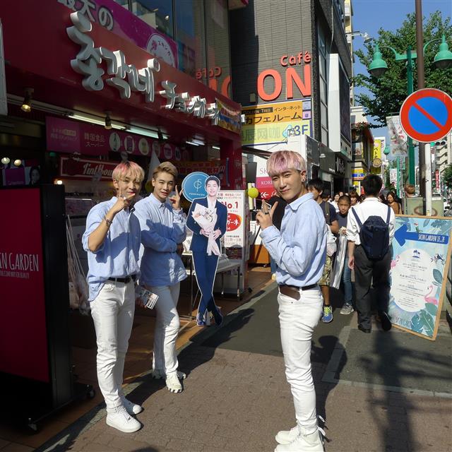 무명의 젊은 한국 남성 보컬 그룹 일원들(염색하고 하늘색 셔츠를 입은 젊은이들)이 신오쿠보 거리에서 자신들의 공연을 알리고 있다.