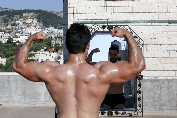 팔레스타인 보디빌딩 트레이너 무하마드 자프레가 10일(현지시간) 웨스트 뱅크 도시 헤브론의 북서쪽에 있는 타르 쿠미야 마을의 코로나19 폐쇄로 체육관이 폐쇄됨에 따라, 학생들과 함께 건물 옥상에서 훈련하는 중 거울을 보며 포즈를 취하고 있다. AFP 연합뉴스