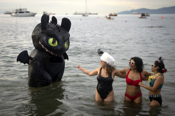 A person wearing a dragon costume stands in the water as three women take a photo during the Polar Bear Swim on New Year‘s Day at English Bay Beach in Vancouver, British Columbia, Monday, Jan. 1, 2024. AP 뉴시스