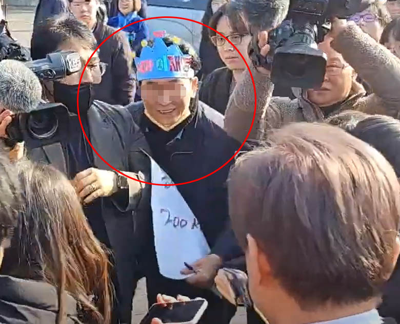 ‘내가 이재명’이라고 적힌 파란색 종이 왕관을 쓴 60대 남성 김모(가운데)씨가 2일 부산 가덕도신공항 부지 현장을 방문한 이재명(오른쪽 뒷모습) 더불어민주당 대표를 향해 미소를 띠며 다가오고 있다. 유튜브 정영일TV 캡처