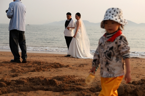 한 중국 어린이가 산둥성 칭다오의 해변에서 결혼 사진을 찍는 신혼 부부 앞에서 모래를 가지고 놀고 있다. 칭다오 로이터 연합뉴스