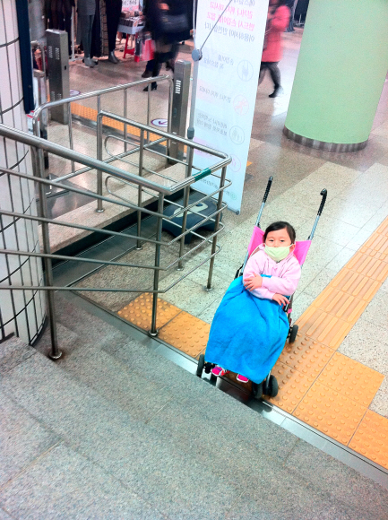 소아암인 신경모세포종을 앓고 있는 6살 수민이가 휠체어 리프트가 고장난 지하철 3호선 고속터미널역 계단 앞에서 유모차에 앉은 채 난감한 표정으로 위쪽을 바라보고 있다. 