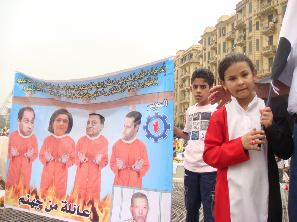 호스니 무바라크 전 이집트 대통령과 가족을 사형대에 세워야 한다는 주장을 담은 플래카드. 그 옆에 이집트 국기 모양의 옷을 입은 소녀가 이집트 국기를 들고 서 있다. 