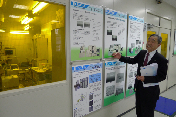 에리오닉스의 세이고 혼메 회장이 하치오지시 본사 복도에 걸려 있는 나노 측정기 설명판을 가리키면서 각 기기의 특징과 기능에 대해 소개하고 있다.