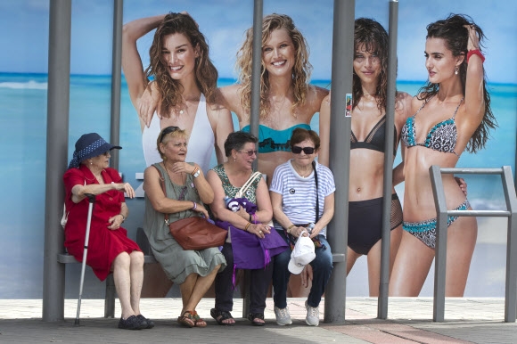 4명의 중년 여성들이 27일(현지시간) 스페인 마드리드에서 수영복을 입고 있는 4명의 젊은 여성들이 나오는 광고 포스터 앞 정류장에서 버스를 기다리고 있다. AP 연합뉴스