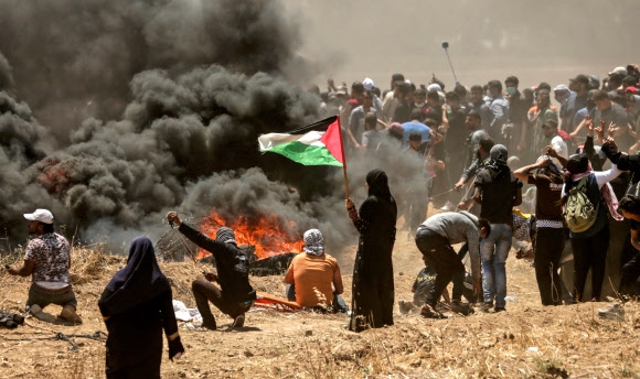 14일(현지시간) 팔레스타인 가자지구에서 한 여성이 타이어를 태운 시커먼 연기를 보며 팔레스타인 국기를 들고 서 있다. 시위대는 이스라엘 주재 미국대사관의 예루살렘 이전에 반대하며 시위를 벌였다. 이스라엘군이 시위대를 향해 발포해 37명이 숨졌다고 가자지구 보건당국은 밝혔다. 2018.5.14 [AFP=연합뉴스]