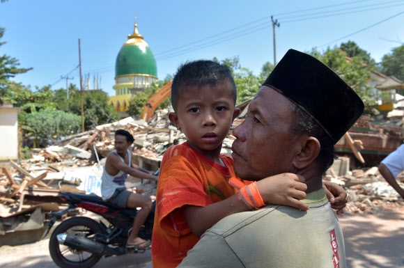 강진으로 대규모 인명피해가 난 인도네시아의 휴양지 롬복 섬에서 한 남성이 무너진 건물 곁에서 아들을 안고 서 있는 모습. AFP 연합뉴스
