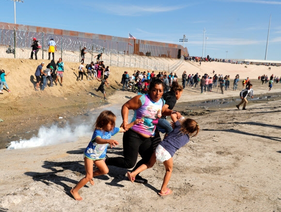 미국 국경수비대원들이 25일(현지시간) 캘리포니아 남부와 국경을 맞댄 멕시코 티후아나의 국경 장벽 너머로 캐러밴에 최루가스를 발사하자 온두라스 출신의 한 어머니가 두 아이를 데리고 달아나고 있다.  티후아나 로이터 연합뉴스