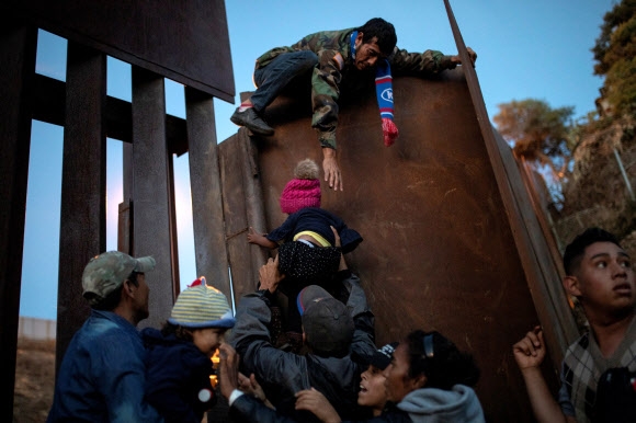 수천㎞를 걸어 미국과 멕시코 국경도시인 티후아나에 도착한 온두라스의 한 남성이 정상적인 방법으로 미국 입국이 어려워지자 2일(현지시간) 불법으로 월경하기 위해 철제 국경벽 중간에 상대적으로 낮은 담벽 위에 올라가 친척들이 받쳐 주는 아이를 잡으려고 필사적으로 팔을 뻗고 있다.  티후아나(멕시코) 로이터 연합뉴스