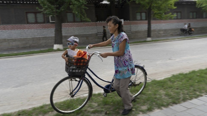선글라스를 폼나게 쓴 어린이가 어머니가 끌고 가는 자전거 앞 좌석에 앉아 있다.