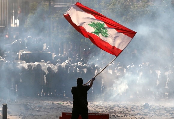 레바논 수도 베이루트에서 8일(현지시간) 벌어진 반정부 시위에서 최루탄 연기가 자욱한 가운데 한 시민이 백향목 문양이 그려진 대형 레바논 국기를 흔들며 시위 진압 경찰과 대치하고 있다. 베이루트 로이터 연합뉴스