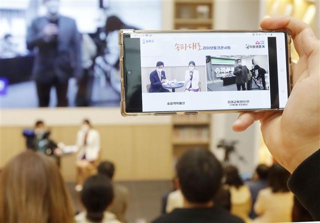 지난 15일 서울 송파구 가락동 책박물관에서 열린 라이브 토크 콘서트 ‘송파대로’에서 한 구민이 원격으로 2원 생중계되는 현장을 스마트폰으로 촬영하고 있다. 박성수(스마트폰 화면 속 왼쪽) 송파구청장이 지역의 대표적인 교육지원 사업인 ‘송파쌤’에 대해 구민들과 이야기하고 있다. 송파구 제공