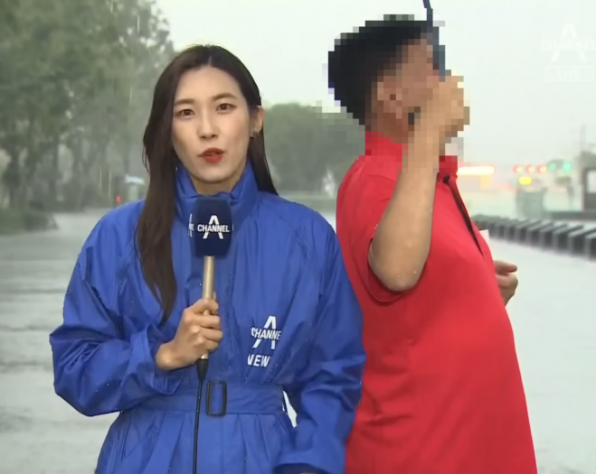 채널A 기자가 비 맞으며 보도하는 가운데 한 시민이 우산을 씌워주고 있다 . 채널A뉴스 캡처
