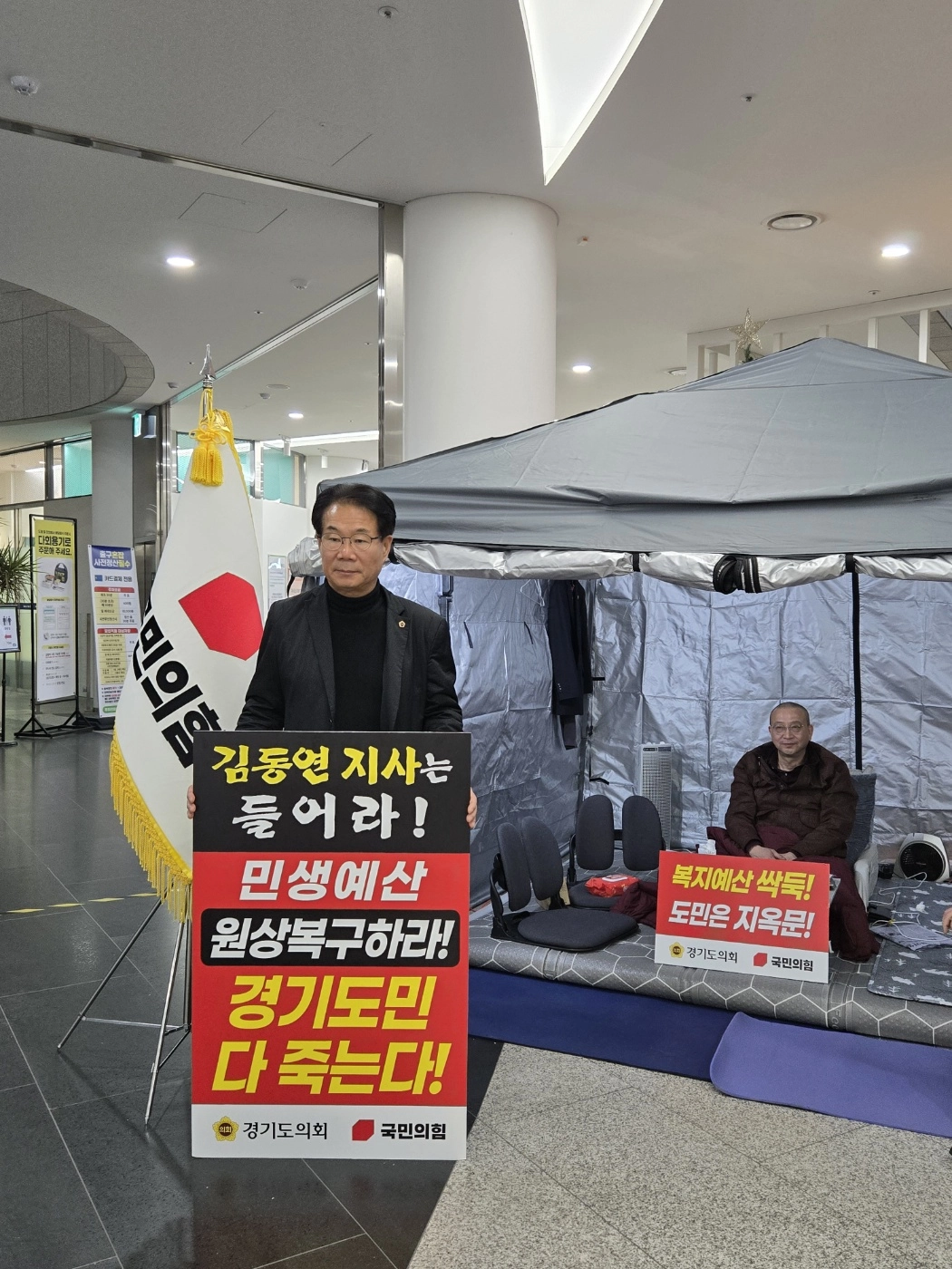 유영두 의원이 도의회 1층 로비 단식농성장 앞에서 손팻말을 들고 이른바 ‘이증도감(李增道減)’식 예산편성으로 인한 도민 피해를 알리며 김동연 도정의 예산 재검토를 촉구하고 있다.(사진=경기도의회)